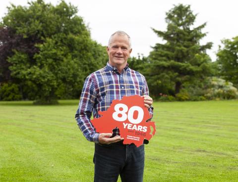 NICHS Chief Executive Declan Cunnane holds a red sign in the shape of Northern Ireland that says 80 years since 1946, thank you for supporting us